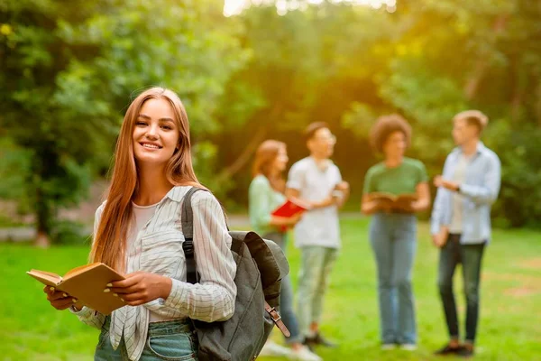 Счастливая улыбающаяся студентка держит книгу, позирующую на открытом воздухе в университетском городке — стоковое фото