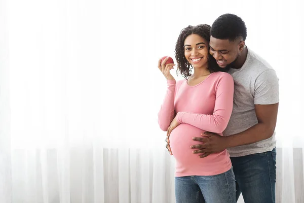 Heureuse femme africaine enceinte mangeant de la pomme pendant que son mari la serre dans ses bras — Photo