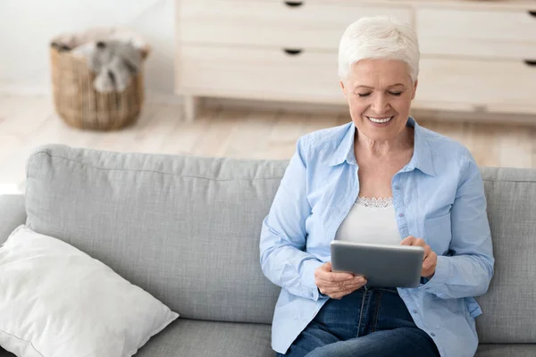 Encantadora senhora idosa sentada no sofá em casa usando computador tablet digital — Fotografia de Stock