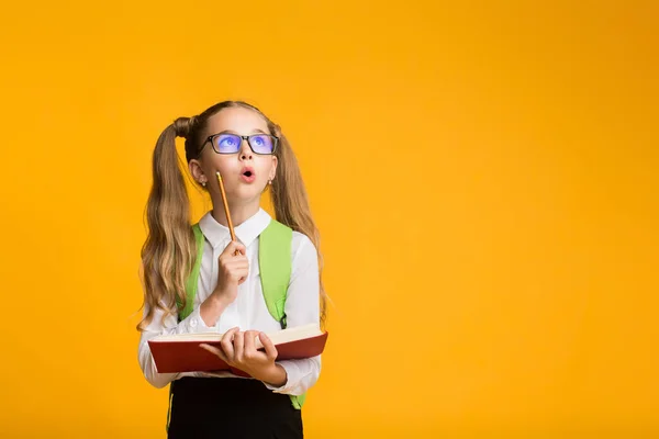 Překvapené školní dívka učení držení knihy a tužka, žluté pozadí — Stock fotografie