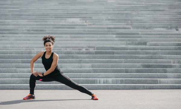 Ejercicios de ajuste después de trotar. Sonriendo chica afroamericana en ropa deportiva haciendo estiramiento para las piernas en el fondo de escaleras grises —  Fotos de Stock