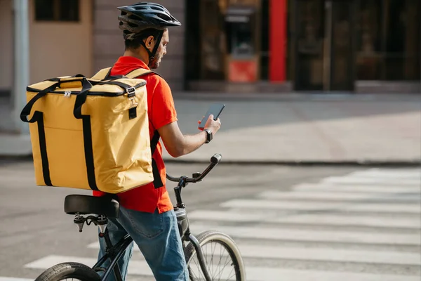 Um entregador de bicicleta. Cara com mochila e capacete parou no cruzamento e olhou para o telefone — Fotografia de Stock