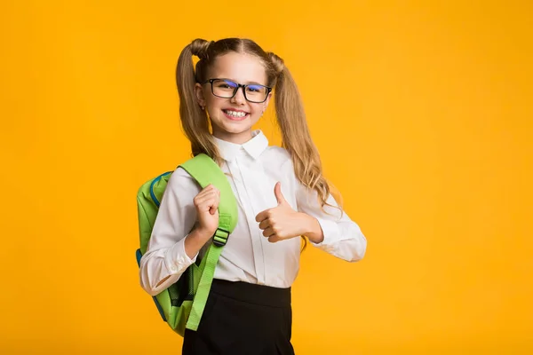 Felice elementare studente ragazza gesturing pollice-up posa su giallo sfondo — Foto Stock