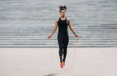 Female athlete does cardio workout. Smiling girl in sportswear with fitness tracker jumping rope clipart