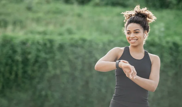 Estilo de vida saludable y control de frecuencia cardíaca. Sonriente chica afroamericana en ropa deportiva con auriculares inalámbricos mirando el rastreador de fitness —  Fotos de Stock