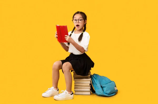 Linda chica de la escuela leyendo libro sentado en los libros, fondo amarillo — Foto de Stock