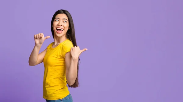 Joyful asian girl pointing on copy space behind her back with thumbs — Stock Photo, Image