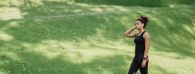 Parkta spor. Kablosuz kulaklıkları ve spor kulaklıkları olan Afro-Amerikalı bir kız parkta yürüyor.