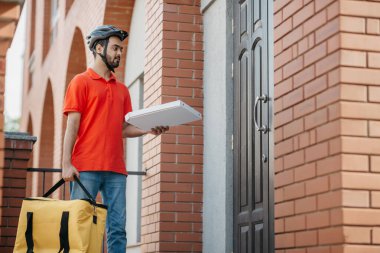 Kurye kapıya pizza getiriyor. Kasklı genç teslimatçı büyük sarı bir çanta ve kutu taşıyor.