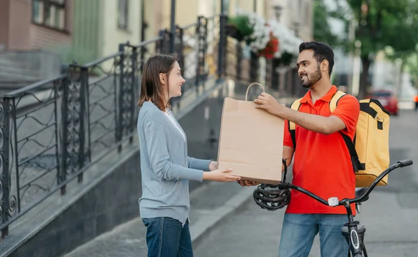 Shopping from home. Friendly courier with backpack and bicycle gives paper bag to client