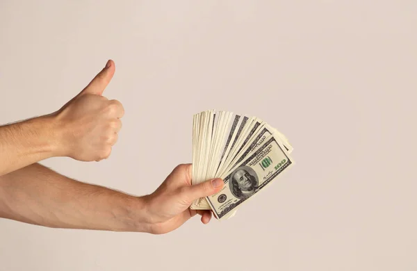 Primer plano del hombre joven sosteniendo ventilador del dinero y mostrando pulgares hacia arriba en el fondo claro, espacio de copia — Foto de Stock