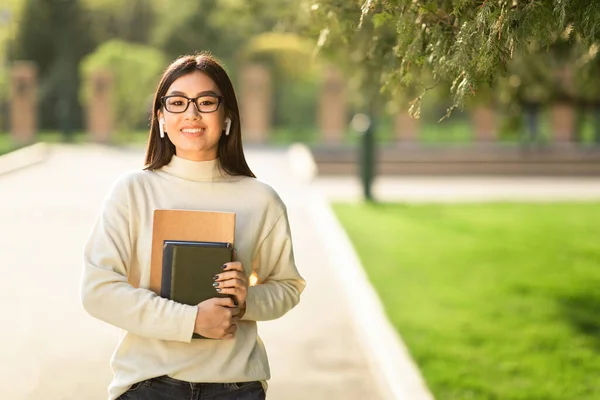 Девушка в очках и наушниках, стоящая в парке, держа книги — стоковое фото