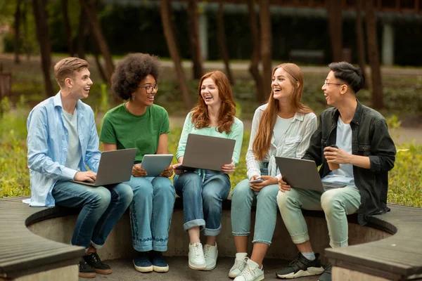 Różnorodni studenci wielokulturowi studiujący na świeżym powietrzu z różnymi urządzeniami elektronicznymi, siedzący na ławce — Zdjęcie stockowe