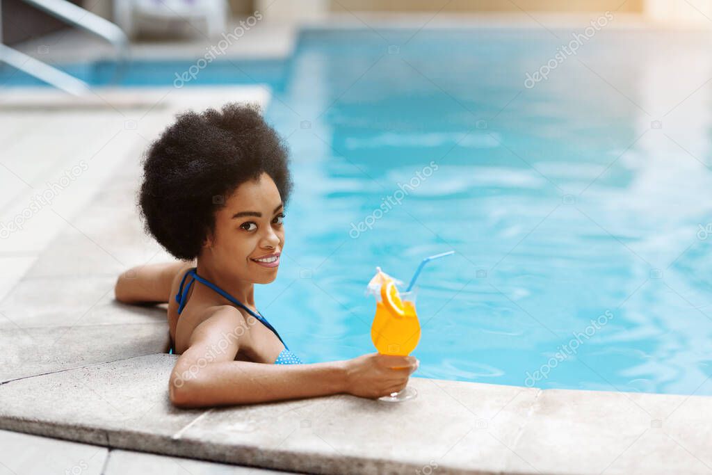 Gorgeous black woman resting by poolside with glass of refreshing cocktail, space for text
