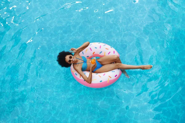 Sexy African American woman with summer drink relaxing on inflatable ring at swimming pool, overhead view