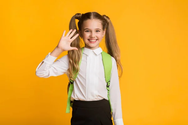 Gelukkig schoolmeisje zwaaien hand glimlachen naar camera op gele achtergrond — Stockfoto