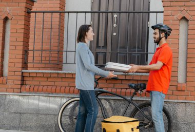 Ekolojik şehir, bisiklet ve teslimat servisi. Kasklı gülen kurye müşteriye pizza veriyor. Girişte, sokakta.