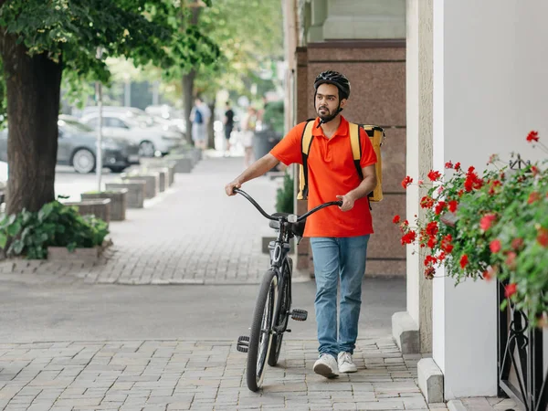 Correio com barba, capacete e saco amarelo, caminha com bicicleta na rua — Fotografia de Stock