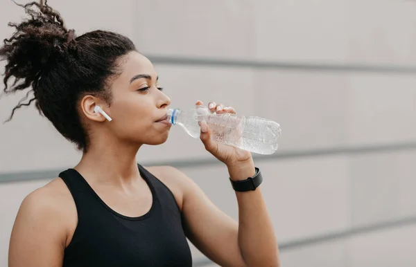 Çapraz, kardiyo eğitimi ve sağlıklı yaşam tarzı. Modern kulaklıklı ve spor takip cihazlı kaslı Afrikalı Amerikalı kız su içiyor. — Stok fotoğraf