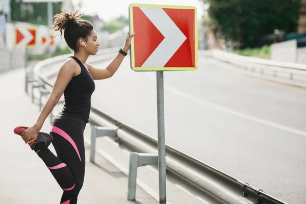 Spor kıyafetli, kablosuz kulaklıklı gülümseyen kız yol tabelasına yaslanıyor ve bacak egzersizleri yapıyor. — Stok fotoğraf