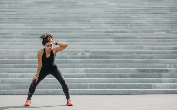 Chica atlética está cansada del entrenamiento activo. Mujer joven afroamericana en ropa deportiva con rastreador de fitness y auriculares inalámbricos descansando en las escaleras de la ciudad — Foto de Stock