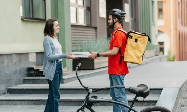 İnternetten yemek siparişi veriyor. Kasklı teslimatçı sarı sırt çantalı ve bisikletli adam müşterilerine evlerinin yakınındaki caddede kutu kutu pizza veriyor.
