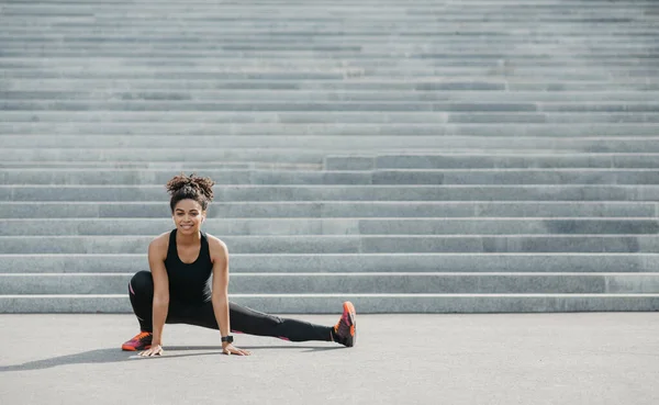 Desporto acessível ao ar livre. Sorrindo mulher afro-americana em sportswear com rastreador de fitness e fones de ouvido fazendo estiramento para perna, perto de escadas da cidade — Fotografia de Stock