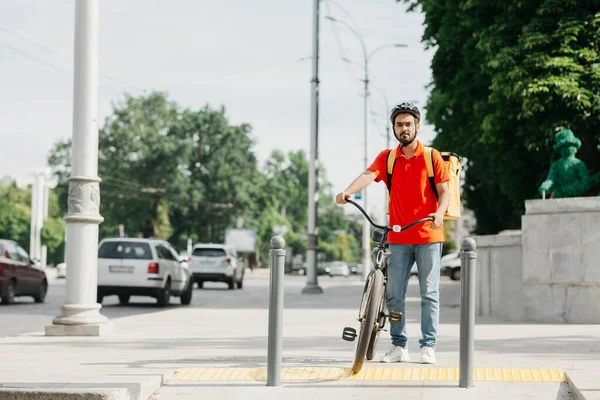 Correio e trânsito municipal. Cara com barba no capacete protetor com mochila amarela e bicicleta, fica na encruzilhada — Fotografia de Stock