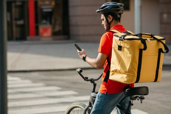 Joven ciclista masculino con bolsa de mensajería utilizando el teléfono móvil en la calle para buscar la dirección del cliente en la aplicación móvil —  Fotos de Stock