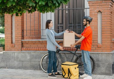 Güvenli bir teslimat. Kasklı gülümseyen kurye, bisikletli büyük sarı sırt çantalı, müşteri için kese kağıdı veriyor.