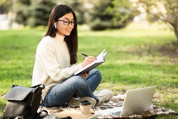Дівчина пише в щоденнику, використовуючи ноутбук, сидячи в парку — стокове фото
