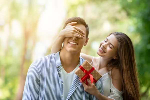 Молодая девушка закрывает глаза своим парням и удивляет его подарком на день рождения в парке — стоковое фото