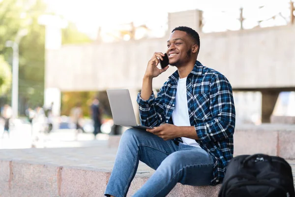 Μαύρος άντρας που χρησιμοποιεί φορητό υπολογιστή, κάθεται στην πόλη με smartphone — Φωτογραφία Αρχείου