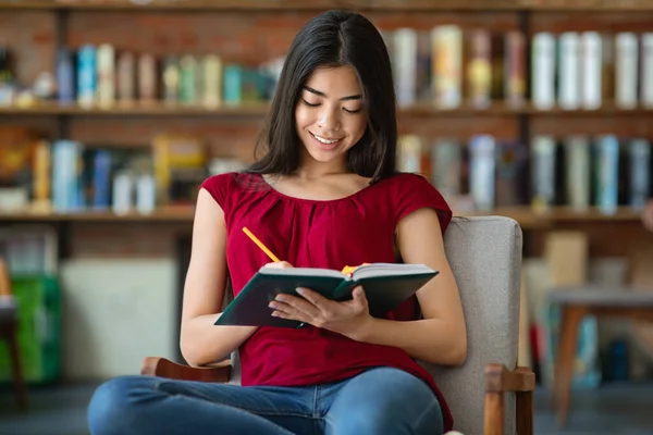 Ázsiai diák lány ül a könyvtárban, és jegyzetel jegyzet — Stock Fotó