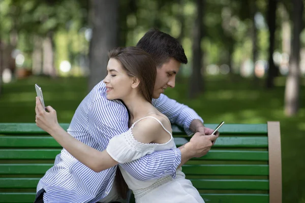 Adicción a Internet. Millennial chico y su novia abrazando y jugando en los teléfonos inteligentes en el parque — Foto de Stock