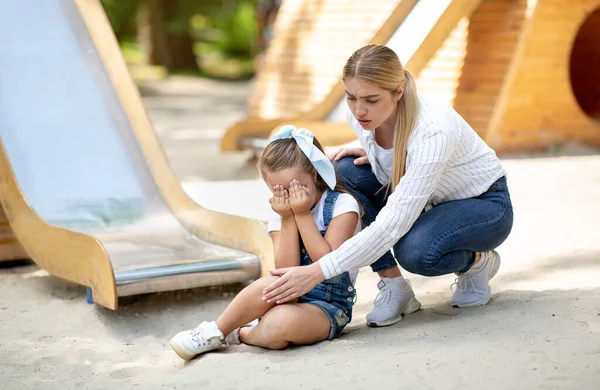 Mãe examinando as filhas do joelho ferido após passeio de corrediça no parque infantil — Fotografia de Stock