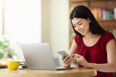 Faydalı bir uygulama. Kafede otururken akıllı telefon ve dizüstü bilgisayar kullanan Asyalı kız.