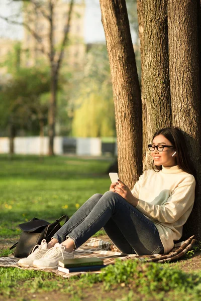 Студентка використовує телефон, сидячи під деревом в парку — стокове фото