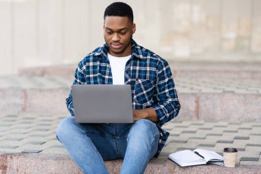 Laptop kullanan bir adam, şehirde basamaklarda oturuyor.