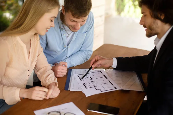 Az ingatlanügynök bemutatja az új otthon tervét a boldog párnak az asztalnál — Stock Fotó
