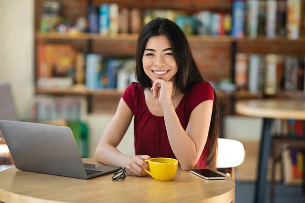 快乐的亚洲女孩在咖啡店的笔记本电脑上边工作边喝咖啡 — 图库照片