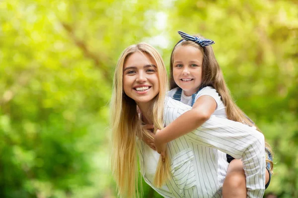 Happy Mother Carrying Daughter Piggyback Outdoors In Park