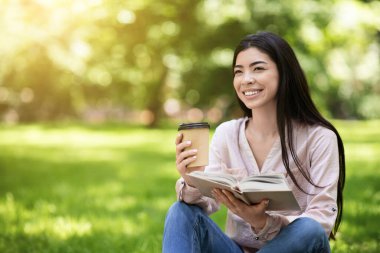 Mutlu Asyalı kız öğrenci parkta kitap okuyor ve kahvenin tadını çıkarıyor.