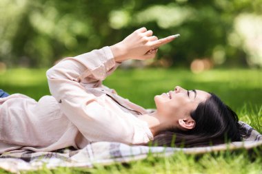 Parkta çim üzerinde akıllı telefonuyla uzanan mutlu Asyalı kız.