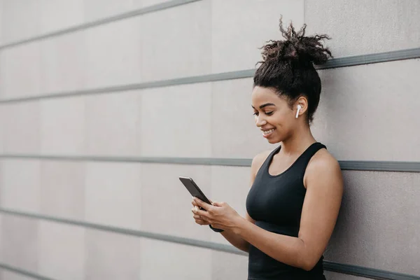 Actividad motivación, vitalidad y deporte. Sonriente chica afroamericana en ropa deportiva con auriculares escribiendo en el teléfono inteligente para el blog de deportes — Foto de Stock