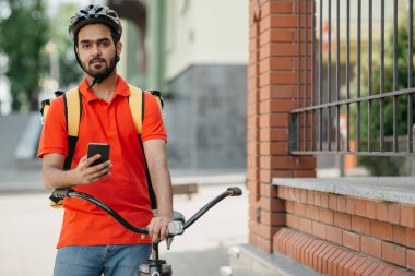 Dost canlısı kurye, sırt çantalı, bisiklete biner ve akıllı telefonu elinde tutar.