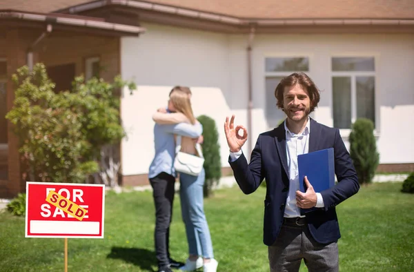 Lächelnder Makler zeigt okay Geste und Millennial-Paar umarmt in der Nähe ihrer neuen Immobilie, Kopierraum — Stockfoto