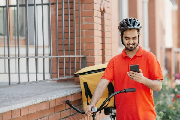 Adres dostawy. Uśmiechnięty kurier w mundurze i kask z plecakiem i oglądaniem rowerów na smartfonie — Zdjęcie stockowe