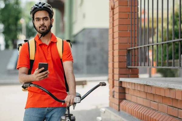 Przyjazny kurier w kasku, z plecakiem, siedzi na rowerze i trzyma smartfona w ręku — Zdjęcie stockowe