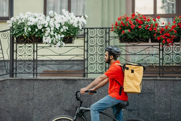 Adam şehirde paket teslimatı yapan bir bisiklete biniyor. Sarı sırt çantalı kurye tarafından çevrimiçi sipariş — Stok fotoğraf